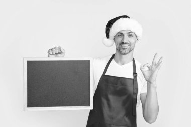 happy mature man in santa hat and apron hold blackboard with copy space. ok. clipart