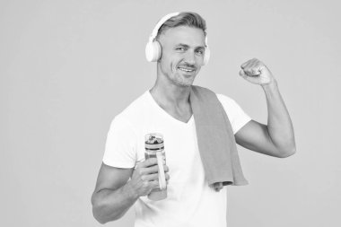 glad fitness man in headphones hold sport bottle. fitness man with sport bottle isolated on yellow. fitness man holding sport bottle in studio. fitness man with towel and bottle after training. clipart