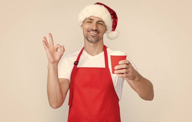 mature christmas man in red santa hat and apron hold coffee cup. ok. clipart