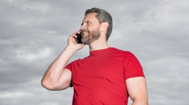 Neşeli adam dışarıda telefonla konuşuyor. Adam telefonda konuşuyor, kırmızı tişört giyiyor. Adamın fotoğrafı telefonda konuşuluyor. Adamın gökyüzü geçmişinde telefon konuşması var..