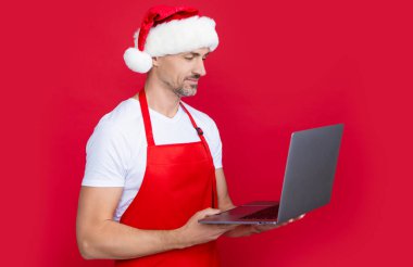 mature man in christmas santa hat and red apron hold computer. clipart