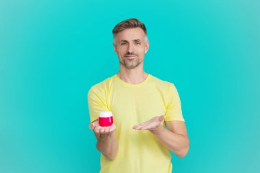 Mavi renkte yüz kremi gösteren bir adam var. Olgun adam cilt kremi kullanır. Günlük alışkanlık ve kişisel bakım. Cilt güzelliği. Erkek güzellik kozmetiği. Erkek kozmetik ürünü sunan adam. Nemlendirici uygula.