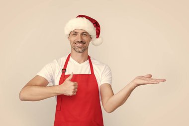 Kırmızı garson önlüğü ve Noel Baba şapkası takan mutlu olgun bir adam. başparmak yukarı.