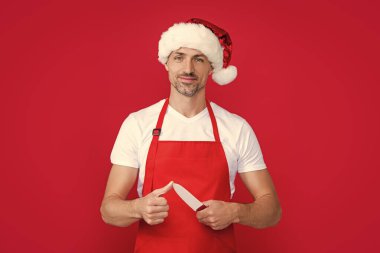 Kırmızı Noel Baba şapkalı ve önlüklü olgun bir adam kırmızı arka planda mutfak bıçağı taşıyor..