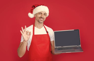 mature man in christmas santa hat and red apron presenting laptop. ok. clipart