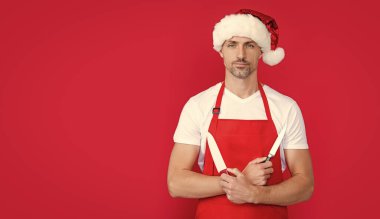 Kırmızı Noel Baba şapkalı ve önlüklü olgun bir adam mutfak bıçağını tutuyor..