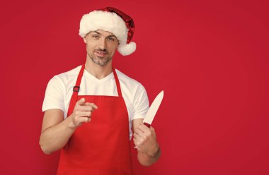 mature christmas man in red santa hat and apron hold kitchen knife. point finger. clipart