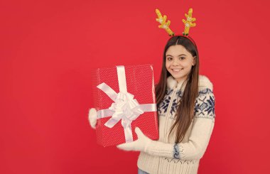 Noel tatili. Geyik boynuzlarıyla yılbaşı hediyesiyle gülümseyen genç kız. Genç kız Noel hediyesi olarak kırmızı arka planda izole edilmiş..