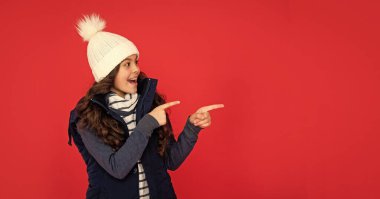 surprised kid with curly hair in hat. teen girl on red background. portrait of child wearing warm clothes. express positive emotion. winter fashion. pointing finger on copy space. clipart