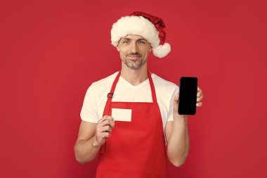 mature man in christmas santa hat and red apron hold smartphone and card. online banking. clipart