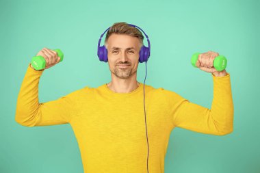 Olgun bir adam mavi üzerine izole edilmiş spor eğitimi yapar. Halterli ve dambılı bir sporcu. Müzikli spor salonu. Spor salonu. Atletik egzersiz için müzik. Güçlü sporcu adam..