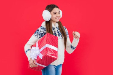 Noel tatili. Kış kulaklıklarıyla noel hediyeli pozitif genç kız. Genç kız Noel hediyesi olarak kırmızı arka planda izole edilmiş..