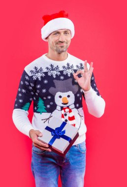 man in xmas santa hat with present box with ok gesture. xmas man isolated on red background. man hold present for xmas in studio. xmas present and man in sweater. clipart