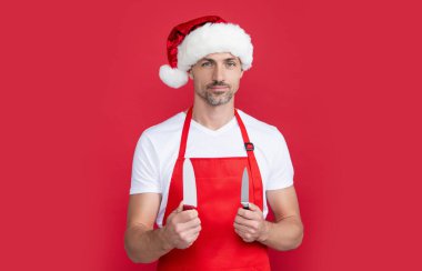 Gülümseyen olgun Noel Baba şapkalı ve önlüklü adam mutfak bıçağını tutuyor..