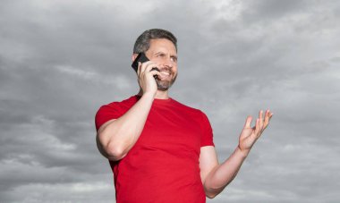 smiling man has phone talk on sky background. man has phone talk outdoor. man has phone talk wear red tshirt. photo of man has phone talk.