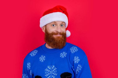 Kafkasyalı mutlu bir adamın portresi Noel süveteri ve kırmızı arka planda izole edilmiş Noel Baba şapkası. Adam Noel 'i kutluyor. Mutlu Noeller..