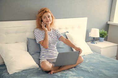Kadın cep telefonuyla konuşuyor ve dizüstü bilgisayarla yatakta otururken gülümsüyor. Mutlu kadın evden telefon ediyor. Telefon görüşmesi.