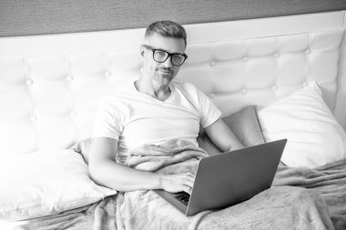 mature man virtual assistant in glasses working on laptop in bed.