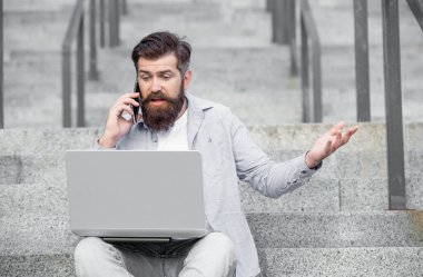 Surprised man talking on mobile phone looking at laptop. Bearded man making mobile call sitting on stairs outdoors. Mobile calling. Mobile communication. Phone conversation.