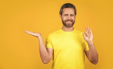 Stüdyoda reklam sunan neşeli adam, tamam. Reklam sunan adamın fotoğrafı. Sarı üzerine izole edilmiş reklam sunan adam. Adam arka planda reklam sunuyor..