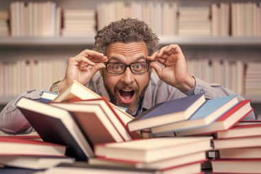 Bir yığın kitabın yanında ev ödevi edebiyatı okuyor. Erkek öğretmen kitap okudu. Üniversitede okul eğitimi. Bilgi günü. Kitap kurdunun fazla çalışması. Okul kütüphanesinde dahi öğretmen. araştırma eğitimi.