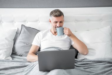 cheerful mature man working on laptop in bed with coffee.