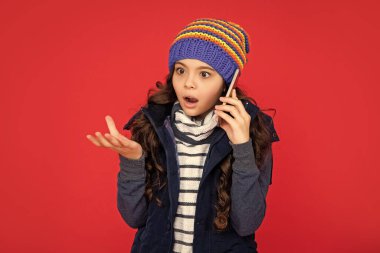 shocked teen girl in down vest with smartphone. portrait of child greeting while phone conversation. communication. kid talking on phone in hat on red background. clipart