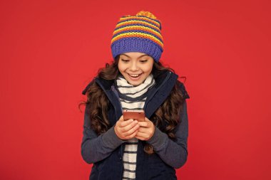 Telefonla blog yazan çocuk portresi. Olumlu duygularını ifade et. Kış tatiliyle selamlıyorum. Gülüşü şaşkın çocuğun kırmızı arka planda şapkayla sohbet edişi. Genç kız akıllı telefondan mesaj atıyor..