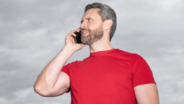Mutlu adamın gökyüzü arka planında telefon konuşması var. Adam dışarıda telefonla konuşuyor. Adam telefonda konuşuyor, kırmızı tişört giyiyor. Bir adamın fotoğrafı telefonda konuşuluyor..