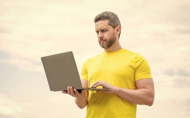 Gökyüzü arka planında dizüstü bilgisayarda çalışan adam.