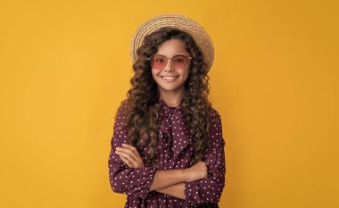 Hasır şapkalı, güneş gözlüklü, uzun, kıvırcık saçlı, sarı arka planda gülümseyen çocuk..