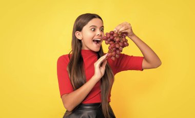 Şaşırmış çocuk sarı arka planda bir demet üzüm tutuyor.