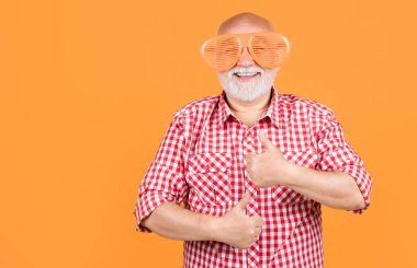 Sarı arka planda damalı gömlekli ve parti gözlüklü neşeli yaşlı bir adam. başparmak yukarı.