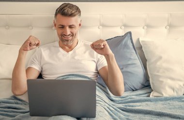 happy mature man working on laptop in bed.