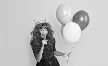 glad child in crown with microphone and party balloon on yellow background.