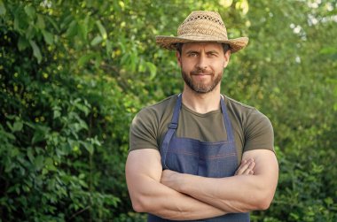 Çiftçi şapkalı, önlüklü, kendine güvenen çiftçi açık havada kollarını kavuşturuyor..