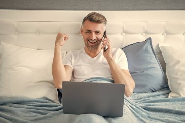 Başarılı bir yetişkin telefonda konuşuyor ve laptopla yatakta çalışıyor..