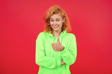 Havalı kapüşonlu giyen kızıl saçlı kız. Pembe üzerine izole edilmiş şık bir kadın. Hareketli moda kız kapüşonlu giyer. Moda kıyafetleri ve tarzı. Kazaklı beyaz bir kadın baş parmağını gösteriyor. Günlük yaşam tarzı.