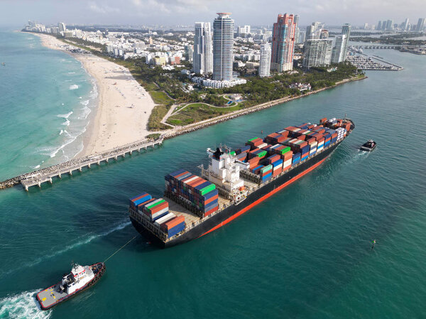 Грузовое грузовое судно. Грузовое судно покидает порт. Контейнерный корабль. Экспорт и импорт. Грузовой бизнес. Порт Майами. Грузовой корабль с контейнером в Майами, вид с воздуха. Массивный стальной корпус.