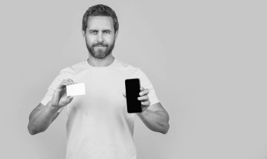 Stüdyoda akıllı telefon ve kart gösteren bir adam, afiş. Akıllı telefon ve kart gösteren bir adamın fotoğrafı. Akıllı telefon ve kart gösteren adam sarıda izole edilmiş. Adam arka planda akıllı telefon ve kart gösteriyor.