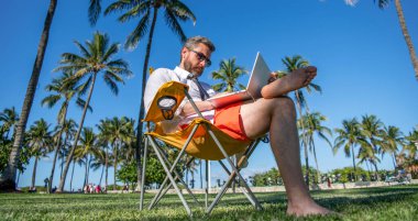 İş gezisi. Plajda serbest çalışan bir adam. Yazın iş adamı. Çevrimiçi serbest çalışma. Dizüstü bilgisayarı olan bir işadamı. Palmiye ağacında dizüstü bilgisayar kullanan bir iş adamı. İş seyahati.