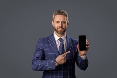 İş uygulamasının reklamı. İspanyol iş adamı telefon ekranını sunuyor. Griler 'de izole edilmiş bir telefon gösteren bir iş adamı. Takım elbiseli adam. Yeni telefon uygulaması. Ürünü sunuyorum. İşaret parmağı.