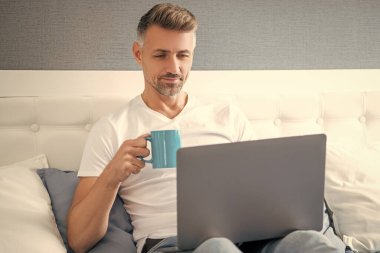 smiling mature man using computer in bed with coffee.