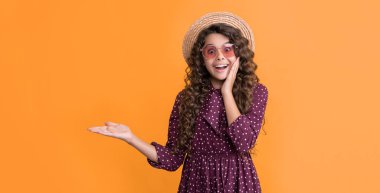 Hasır şapkalı ve güneş gözlüklü uzun kıvırcık saçlı çocuk ürününü sunuyor..