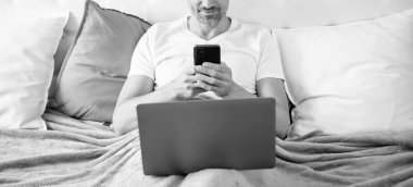 Neşeli, olgun bir adam telefonda konuşuyor ve laptopla yatakta çalışıyor..