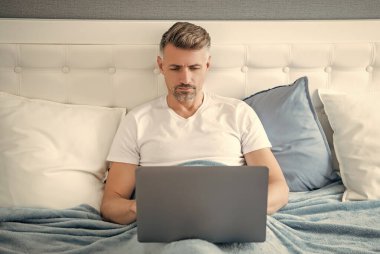 concentrated mature man working on laptop in bed.