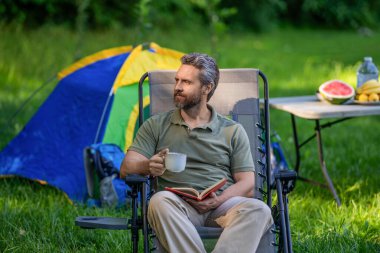 Adam ormanda sabah kampı kahvesi içiyor. Yürüyüşçü adam dışarıda rahatlar. Yaz kampı ve doğa tatili. Adam kamp hakkında kitap okudu. Kamp macerası. Yürüyüş konsepti. Kamp çadırı. Kahve ve kitap..