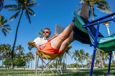 Palmiye ağacında dizüstü bilgisayar kullanan bir iş adamı. İş gezisi. Plajda serbest çalışan bir adam. Yazın iş adamı. Çevrimiçi serbest çalışma. Dizüstü bilgisayarı olan bir işadamı. Şirket seyahat planı.