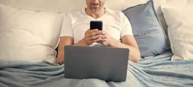 Neşeli, olgun bir adam telefonda konuşuyor ve laptopla yatakta çalışıyor..