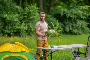 Kamp ve doğa pikniği. Adam kampı. Karpuz kampı pikniği. Lezzetli sağlıklı yiyecekler. Kampta taze yiyecek. Yaz kampında elinde karpuz tutan adam. Yaz günü erkek yürüyüşçüsü. Meyve yiyin..